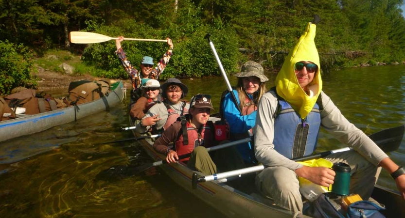 A group of people wearing lifejackets sit in a canoe. One person is wearing a banana costume, 